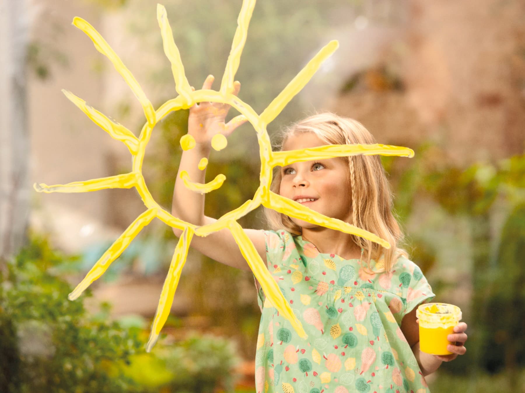 Ein Mädchen im hellgrünen Kleid malt lachend eine Sonne mit gelber Fingerfarbe auf ein Fenster