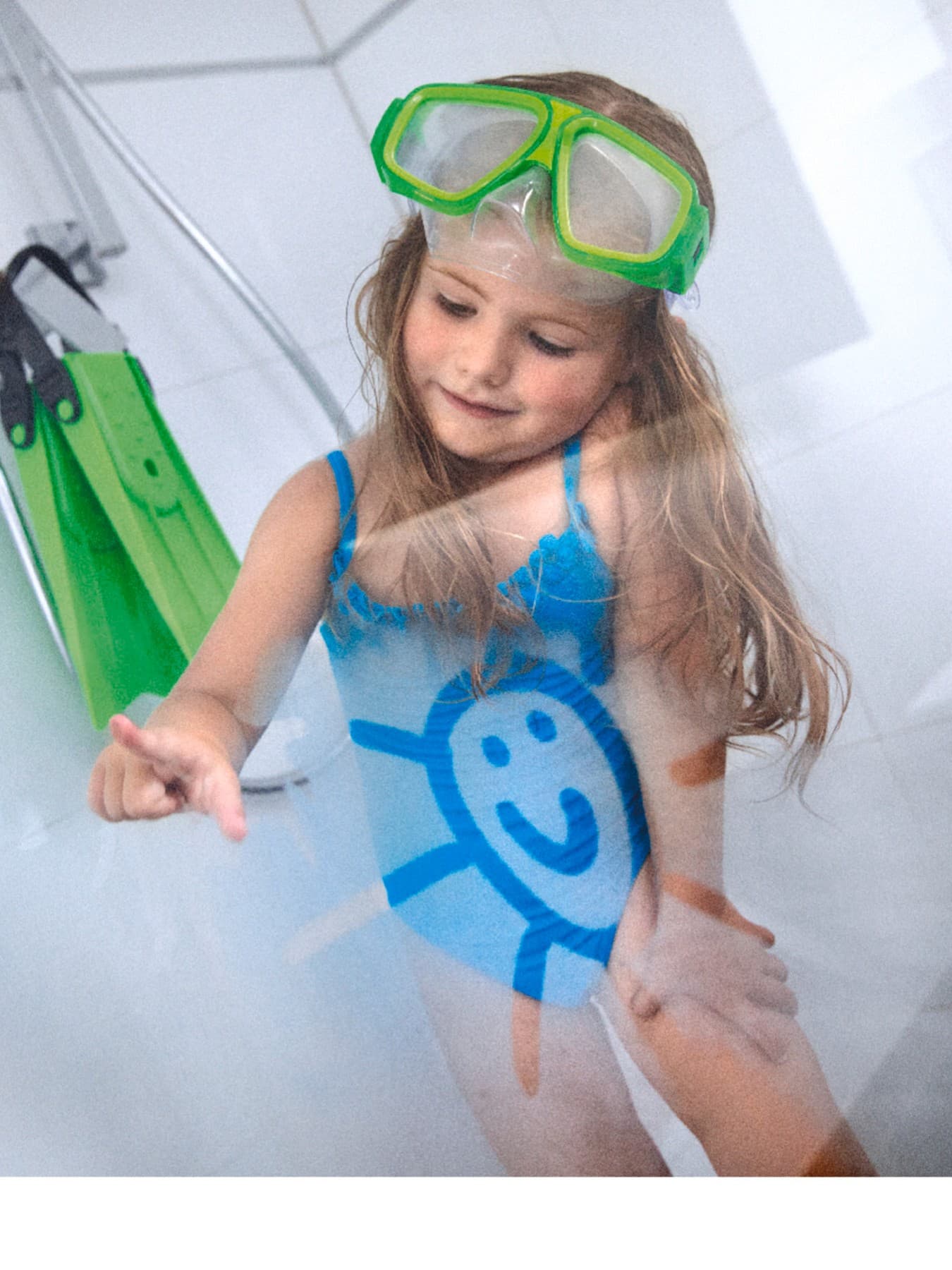 Ein Mädchen im blauen Badeanzug mit Taucherbrille und Flossen in einer Dusche mit beschlagener Duschwand. Auf die Duschwand malt sie eine lachende Sonne.