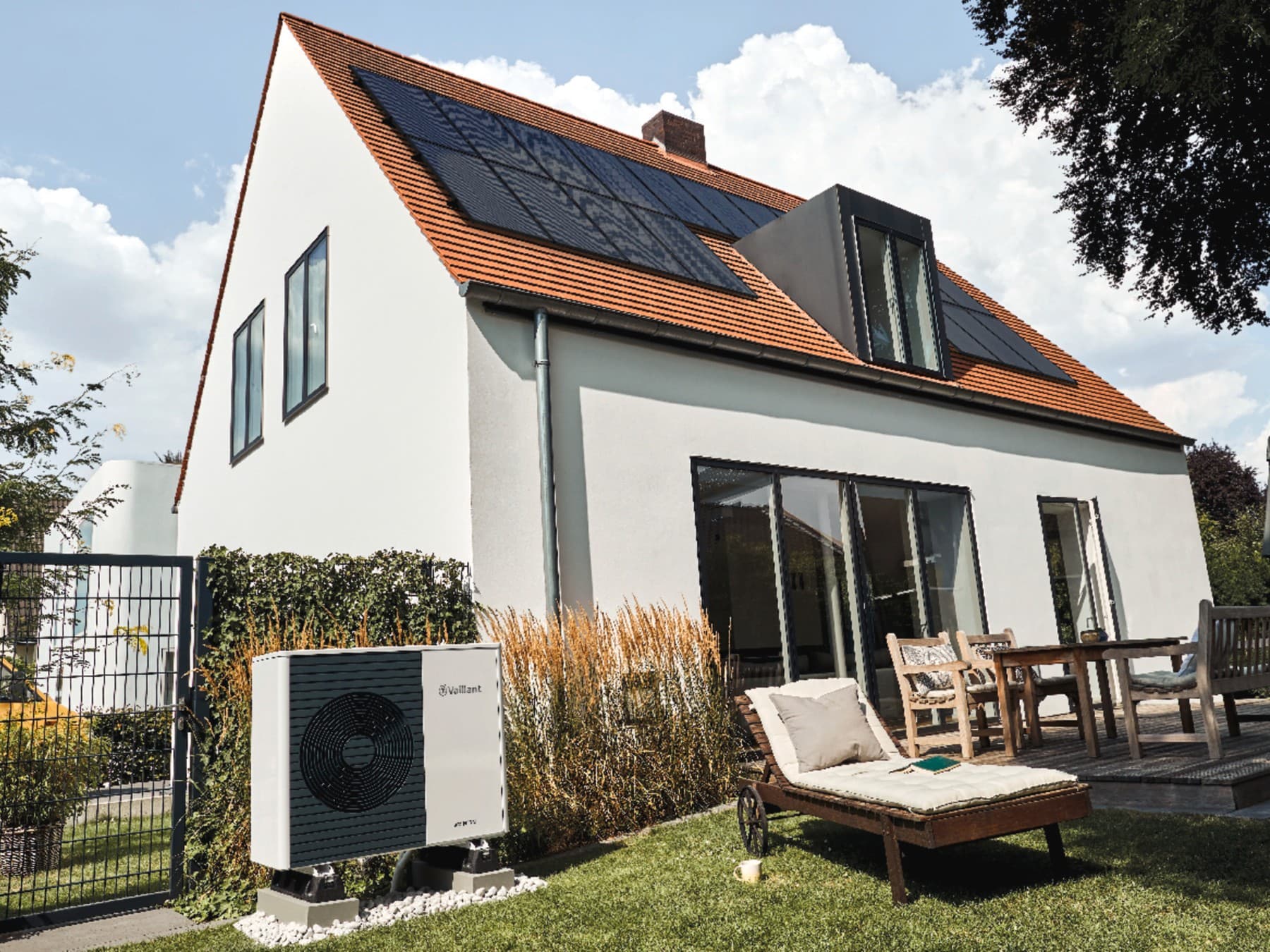 Ein Haus vom Garten aus fotografiert mit Solarpaneelen auf dem Dach und einer Wärmepumpe neben dem Gartentor.