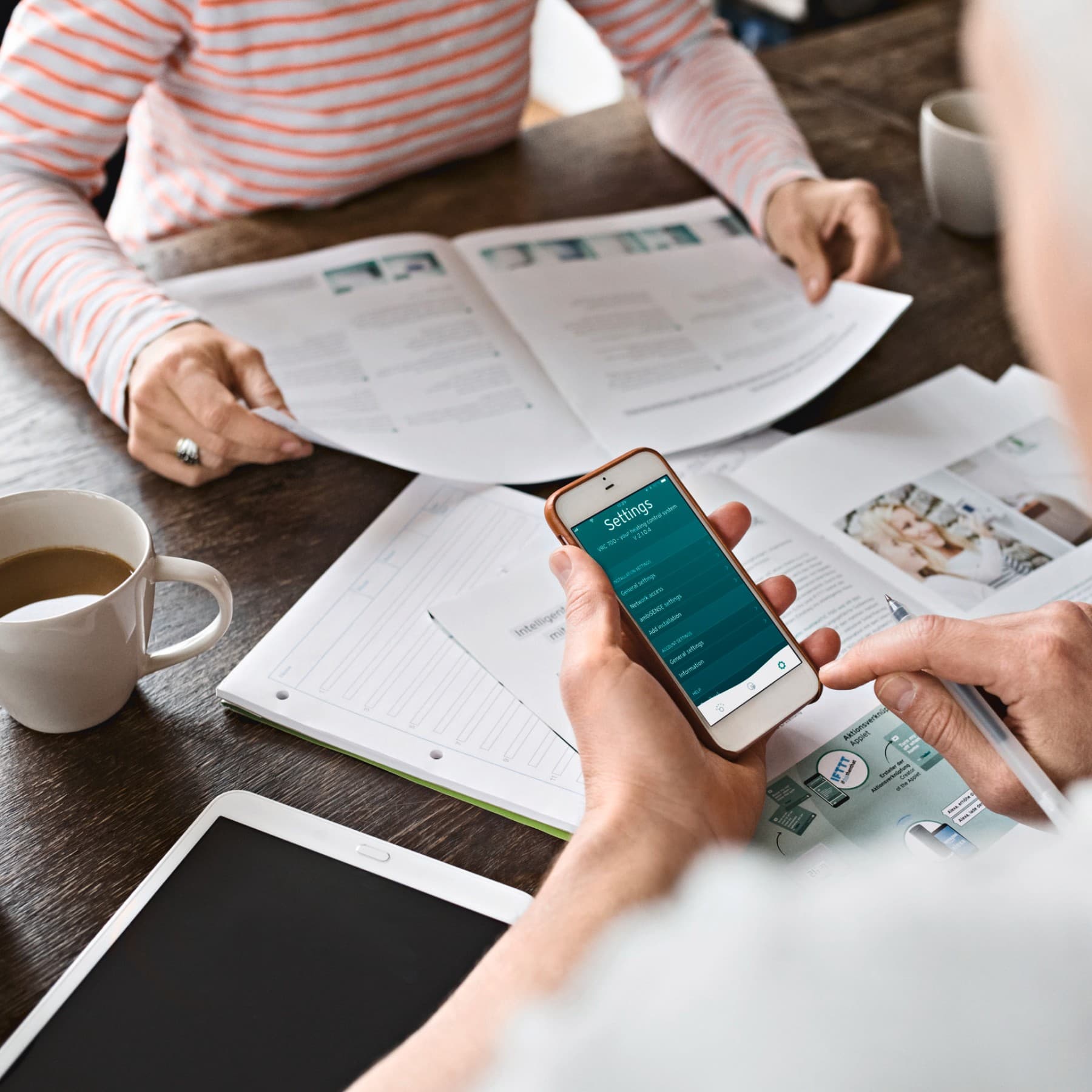 Zwei Menschen sitzen sich an einem Tisch gegenüber, man schaut über die Schulter eines Mannes, der ein Smartphone in der Hand hält. Auf dem Tisch liegen zahlreiche Unterlagen und Dokumente.