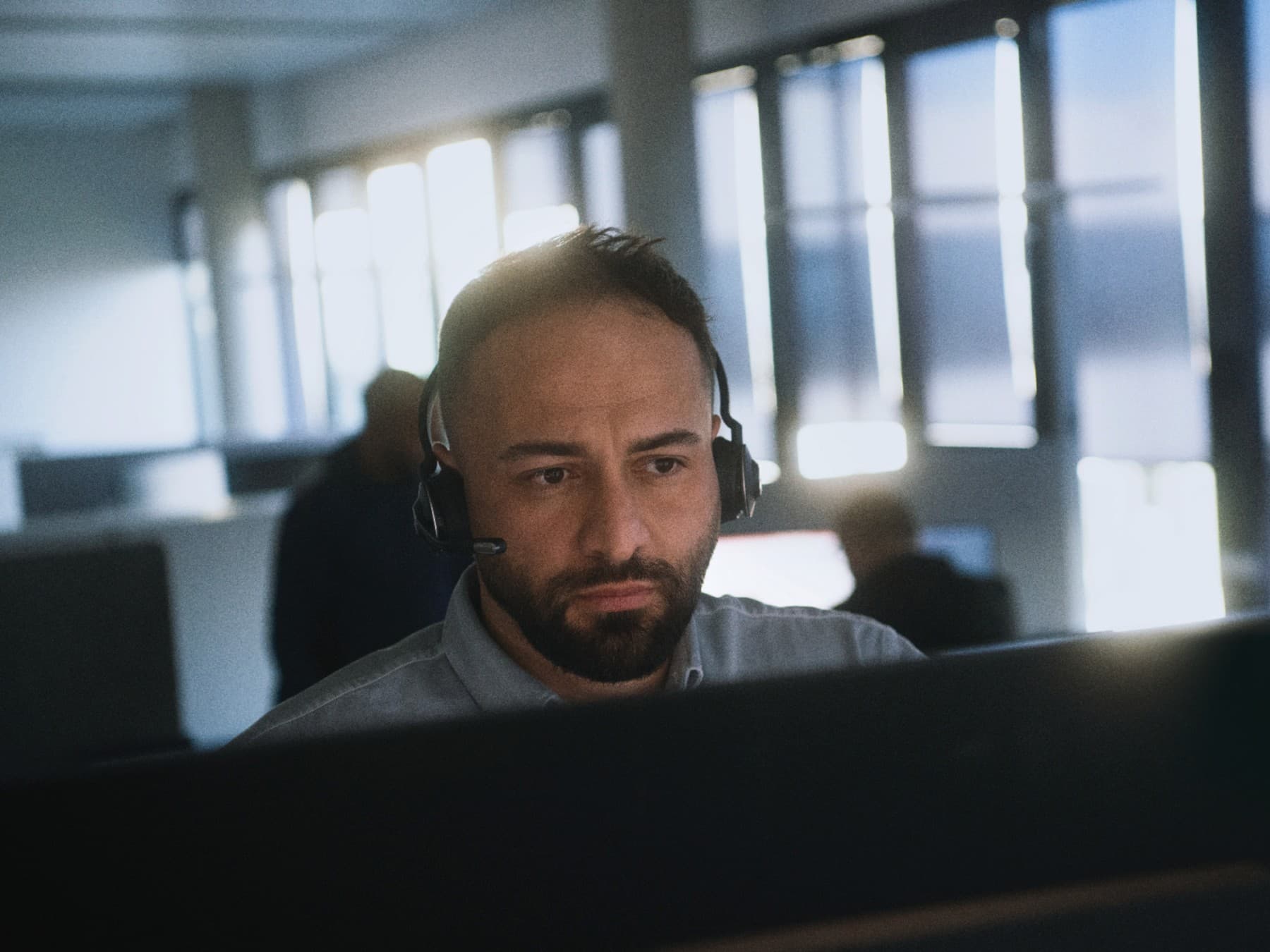 Mann mit Headset sitzt konzentriert vor einem Computerbildschirm in einem Büro mit mehreren Arbeitsplätzen im Hintergrund.