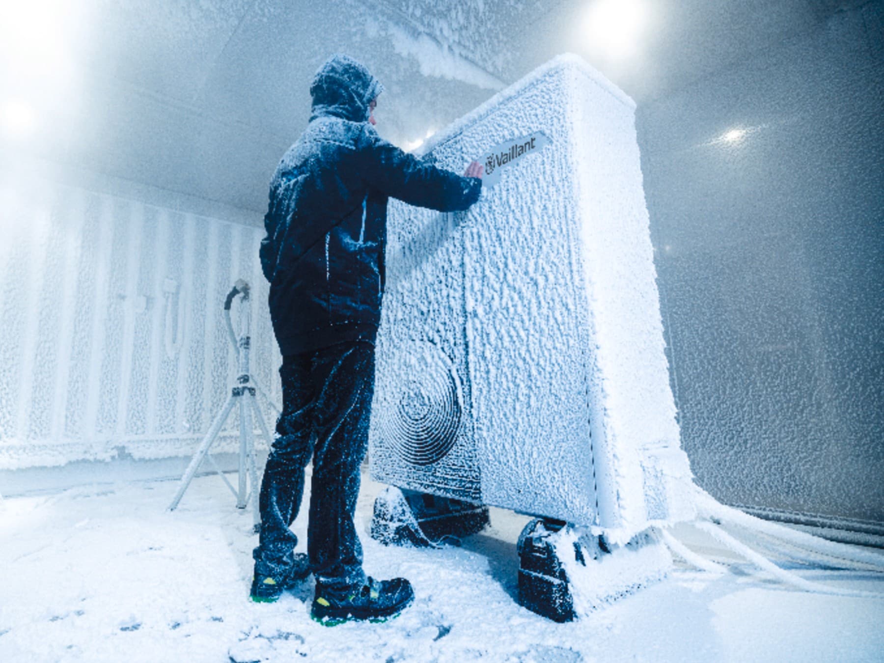 Eine Vaillant Wärmepumpe steht in einem kalten Testraum, in dem es schneit und friert. Ein Mitarbeiter steht davor und entfernt den Schnee vom Vaillant Logo.