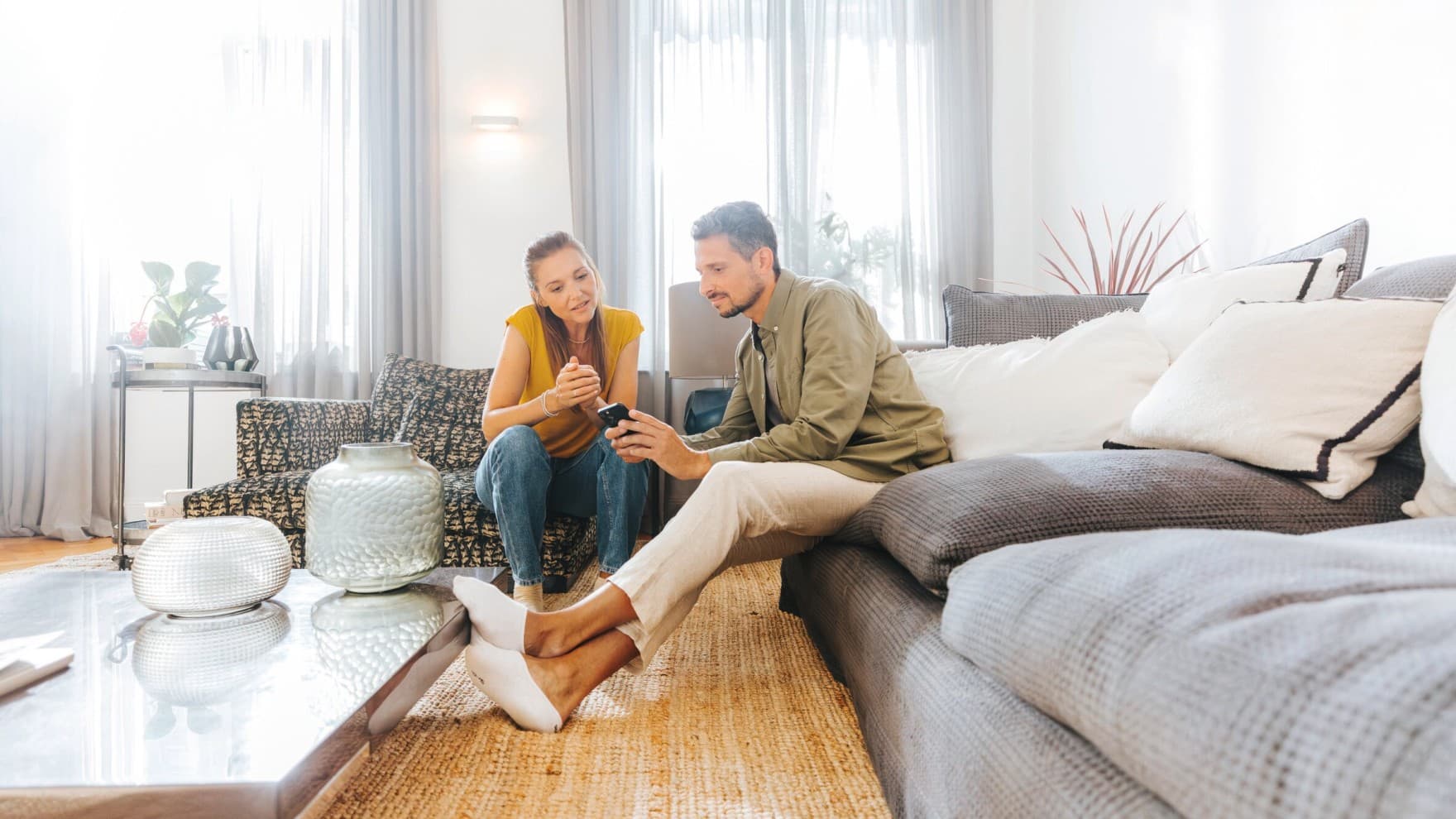 Eine Frau und ein Mann sitzen auf dem Sofa im Wohnzimmer und bedienen den Vaillant-Heizungsregler auf dem Smartphone in der Hand