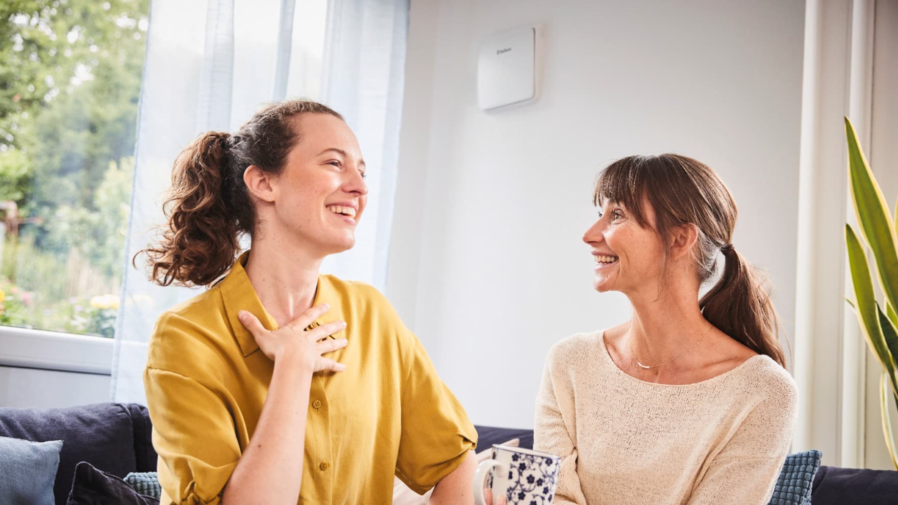 Zwei Frauen auf einer Couch. Im Hintergrund ist an der Wand eine Lüftung zu sehen.