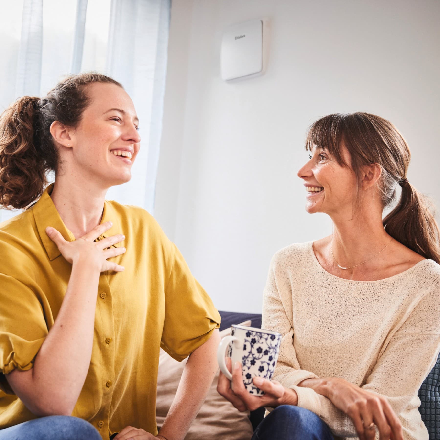 Zwei lachende Frauen sitzen im Wohnzimmer auf der Couch, an der Wand ist die dezente Wohnraumlüftung sichtbar.