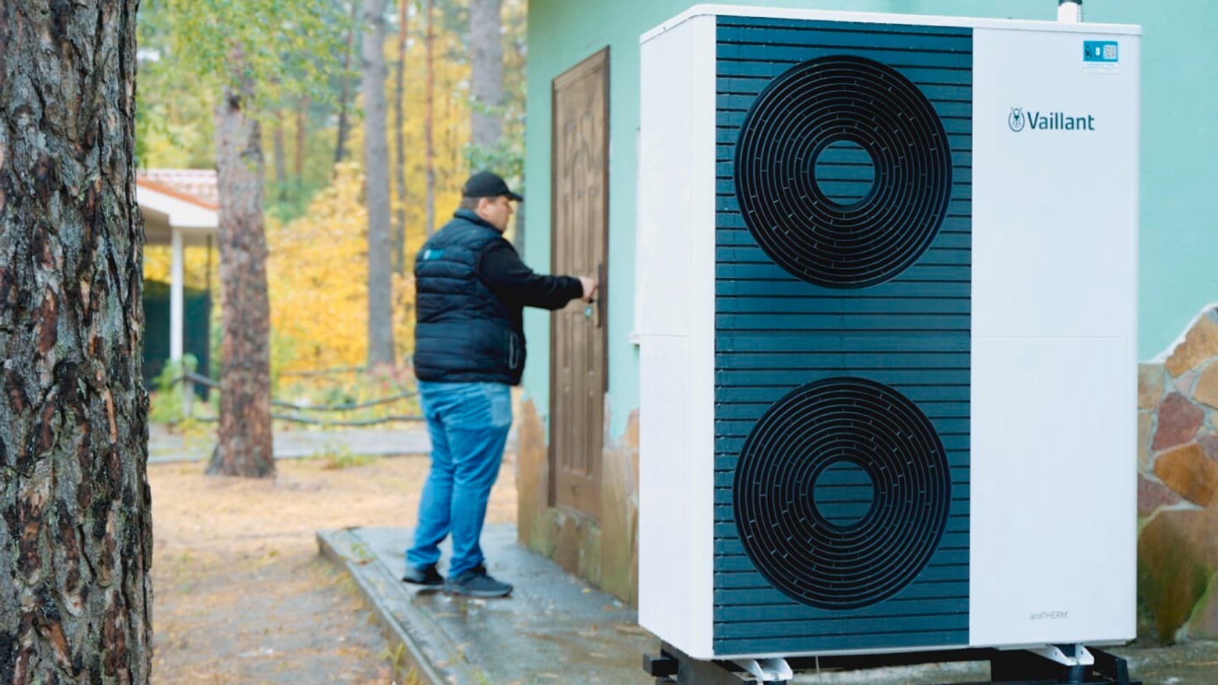 Eine Vaillant Wärmepumpe mit zwei Ventilatoren steht vor einem Haus im SOS Kinderdorf Brovary. Neben dem Haus ist ein Weg mit mehreren Bäumen, im Hintergrund steht ein Mann an der Tür des Hauses und hält seine Hand auf die Klinke.