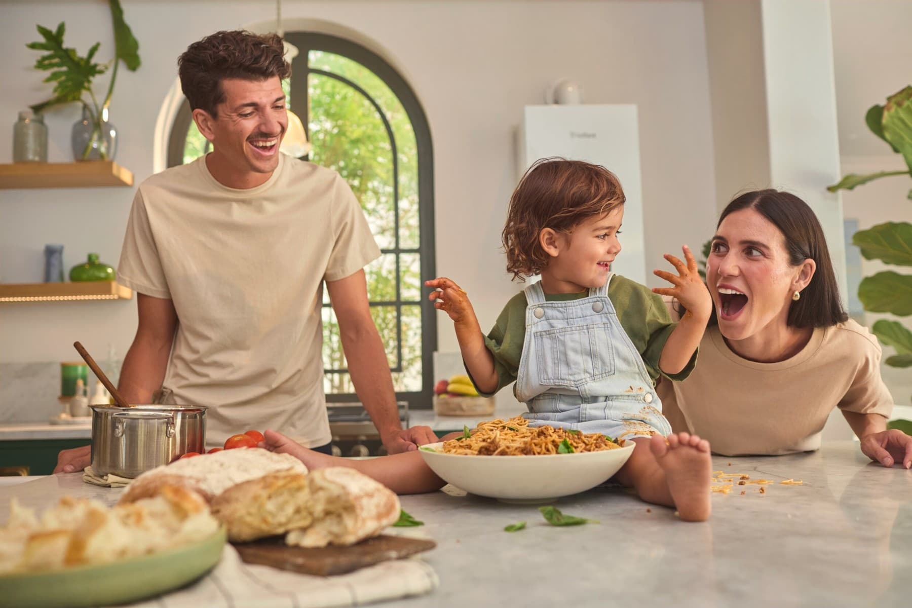 Ein Mann und eine Frau stehen lachend und herumalbernd in einer Küche an einer Kochinsel. Im Hintergrund sieht man Pflanzen auf Regalen, ein Rundbogenfenster und eine Vaillant Gasheizung an der Wand.