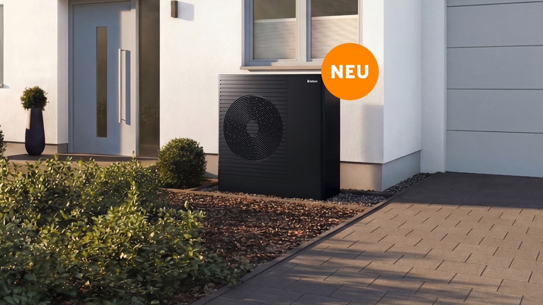 Die Vaillant aroTHERM pro steht vor einem Haus direkt unter dem Fenster rechts neben der Eingangstür. An der Wärmepumpe ist ein orangefarbener Störer "NEU"